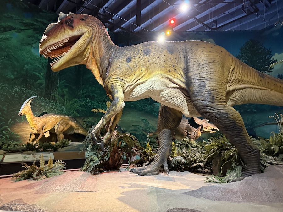 Animatronic T. rex roaring in a prehistoric jungle scene.