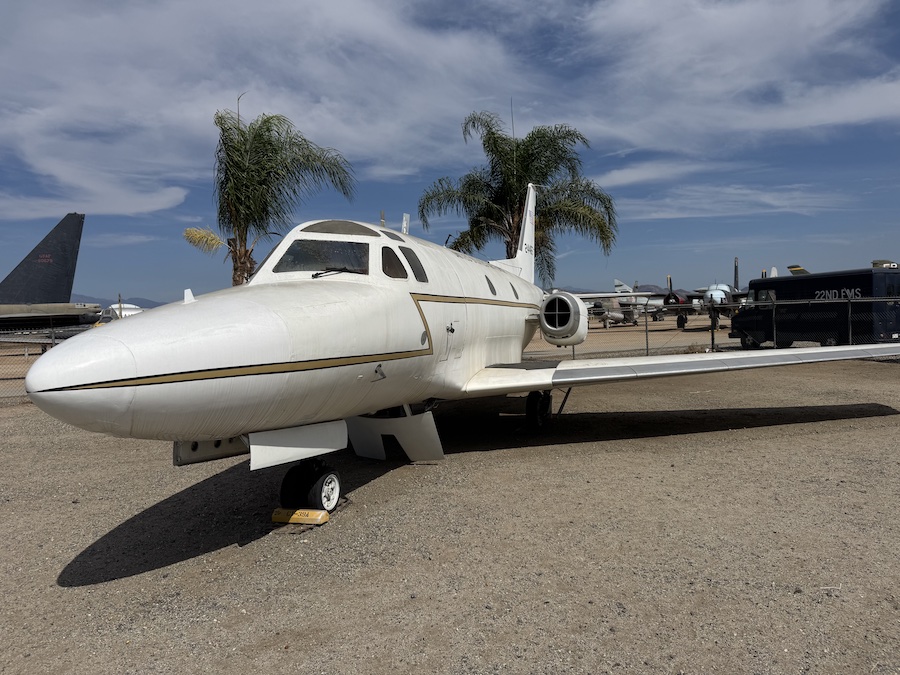 A small, sleek white jet with gold stripes is parked outdoors, with two palm trees behind it.