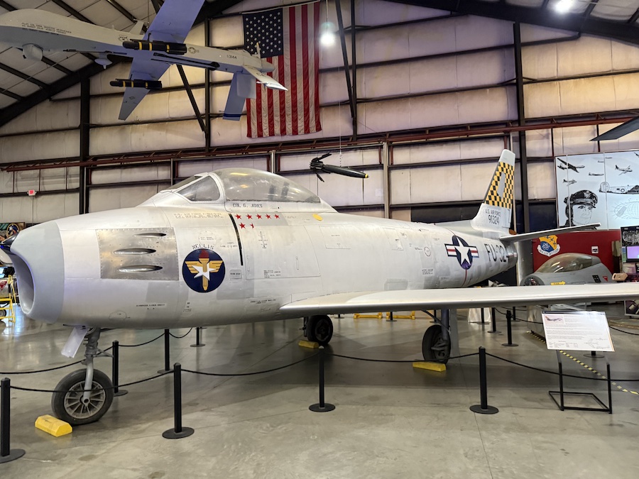 A polished, silver North American F-86F Sabre jet is displayed indoors, roped off for visitors.