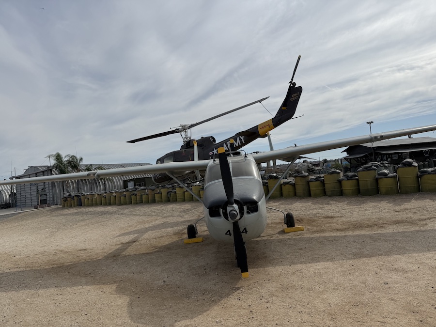 An Army-marked Cessna L-19 Bird Dog plane with a prominent front propeller is parked on a dirt lot.