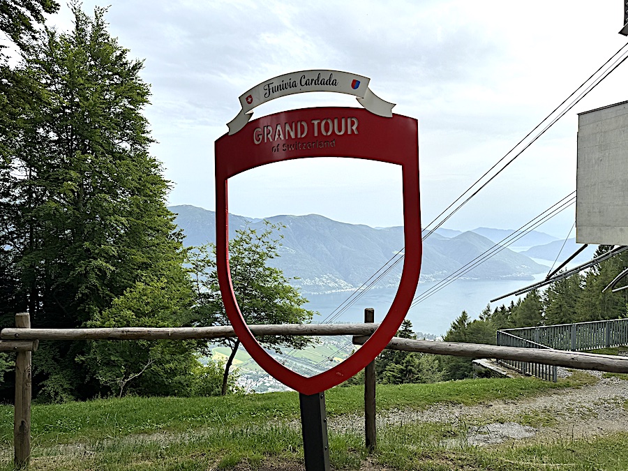 The Grand Tour of Switzerland sign framing a stunning view of the vast lake and the surrounding green mountains and valleys.