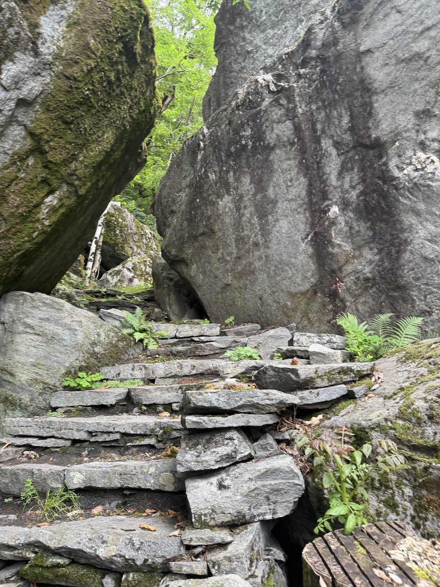 A rugged, natural stone staircase is built into a path between two large, grey moss-covered boulders.