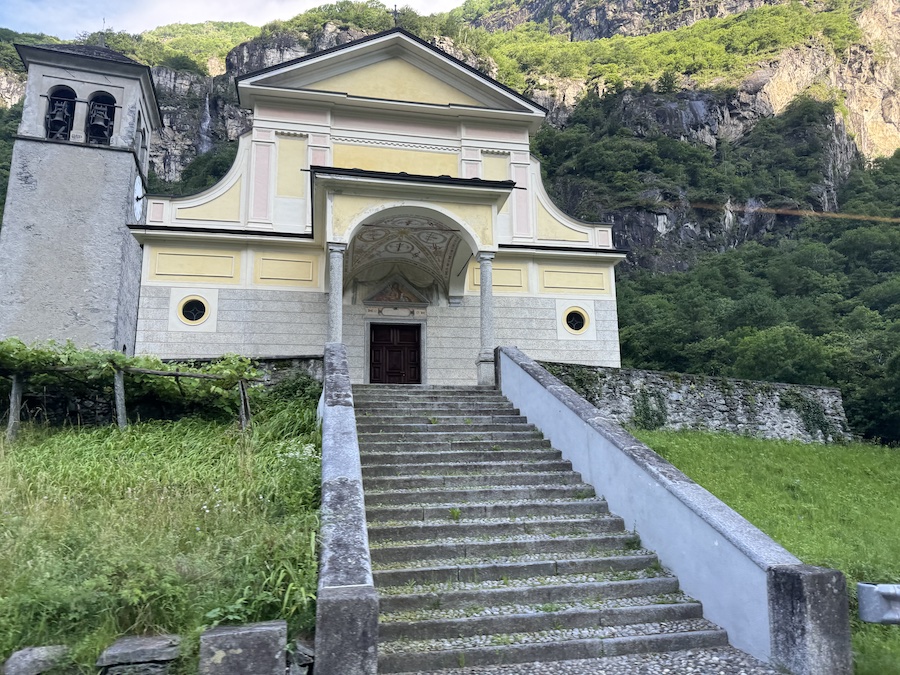 Exterior view of the Church of Santa Maria Assunta and San Giovanni Battista in Cevio, surrounded by mountains.