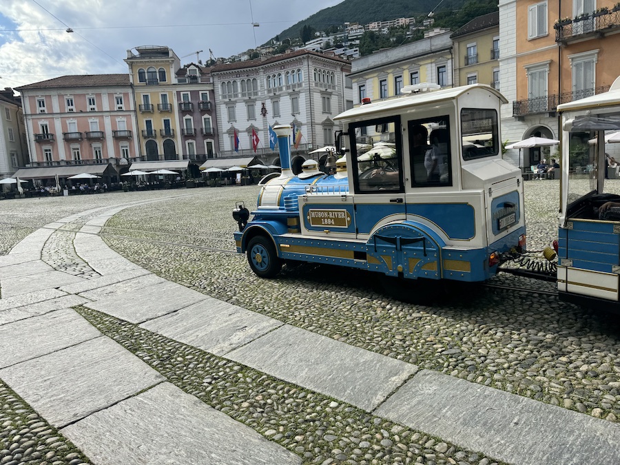 A small tourist train sits on a large cobblestone plaza in Locarno.