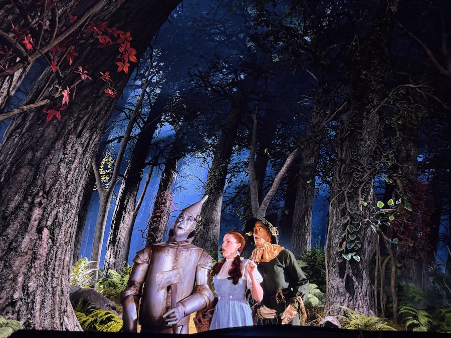 Digital screen display showing Dorothy, the Tin Man, and the Scarecrow standing together in a dark, ominous forest.