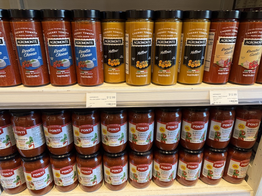 Colorful glass jars of Agromonte and Ponti pasta sauces displayed neatly on a wooden market shelf.