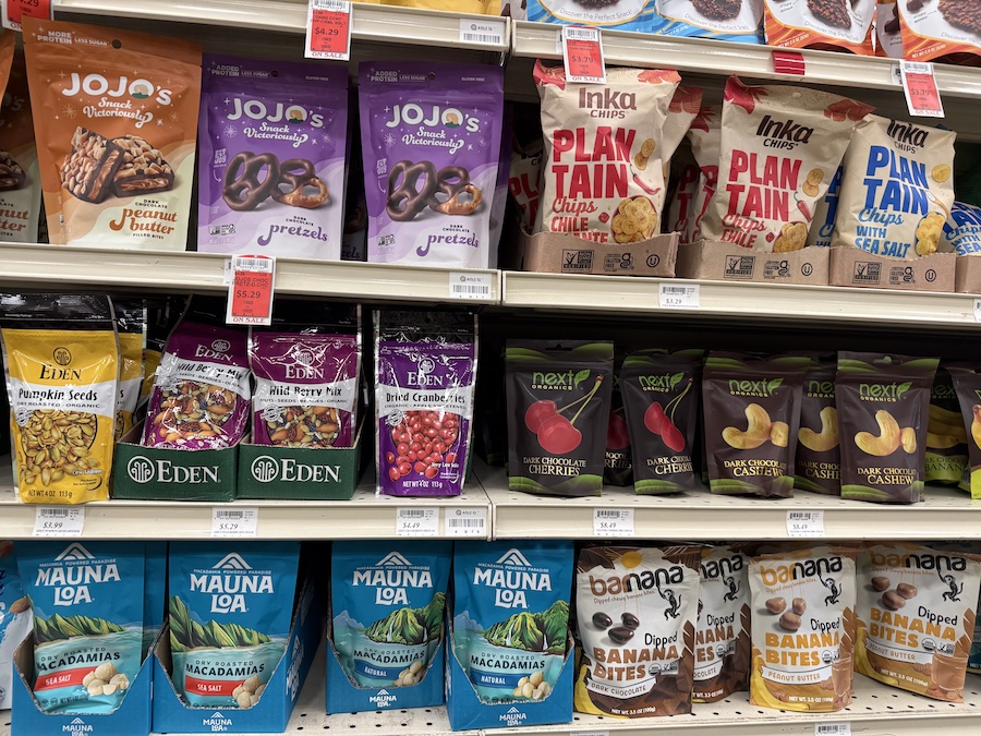 An assortment of roasted nuts and dried fruits from Eden, Next Organics, and Mauna Loa arranged on wooden shelves.