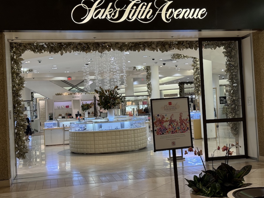The Saks Fifth Avenue storefront inside South Coast Plaza, elegantly lit with window displays showcasing high-end fashion and accessories.