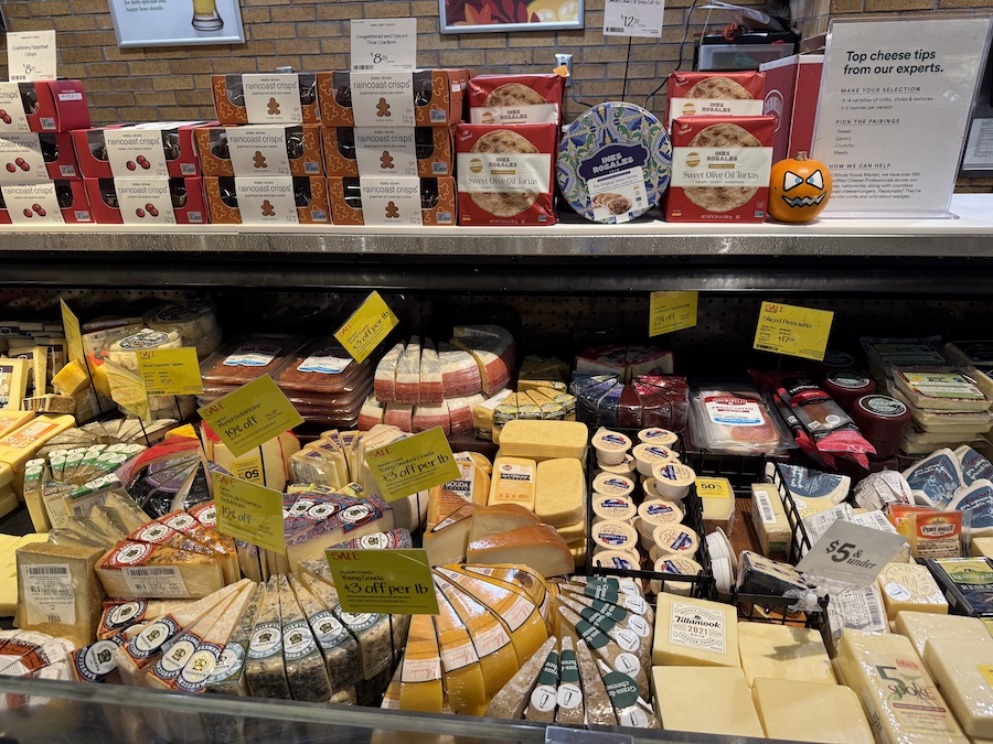A colorful cheese display at Whole Foods featuring artisanal blocks and wheels of cheddar, brie, gouda, and goat cheese from local and international brands.