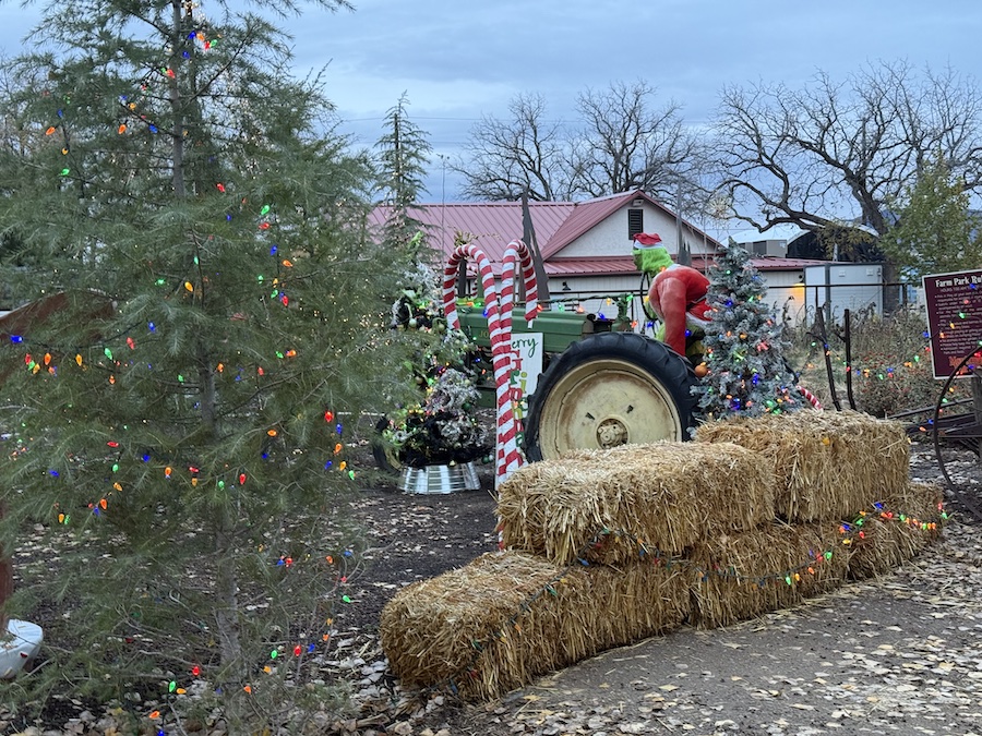 A humorous scene of the Grinch sitting on an old green John Deere tractor, decorated for Christmas with lights and festive accents.