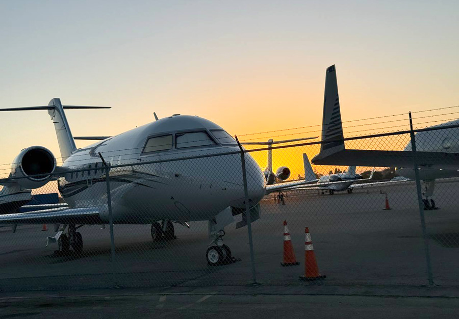 Several business jets parked behind an airport fence at sunset